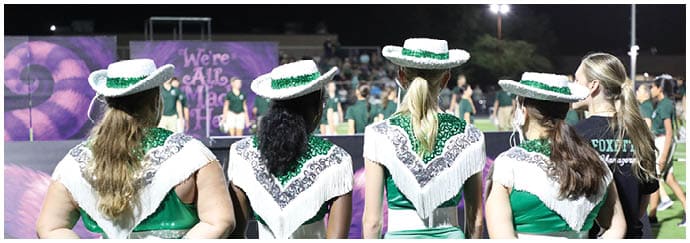 Four people wearing green and white dance team uniforms and hats stand with their backs to the camera on a sports field while other people are visible in the background. Descripci n generada con IA