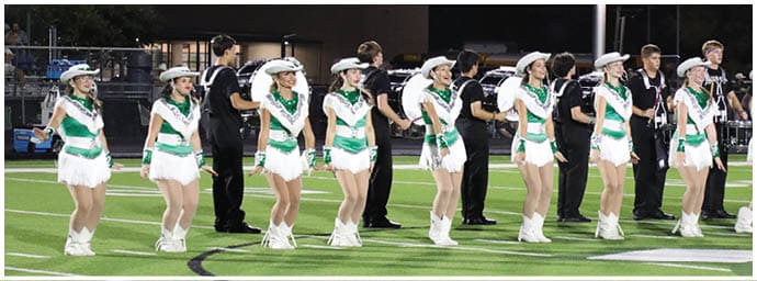 A group of people dressed in matching uniforms and hats are standing in a line outdoors at night, possibly preparing for a performance or event. Descripci n generada con IA
