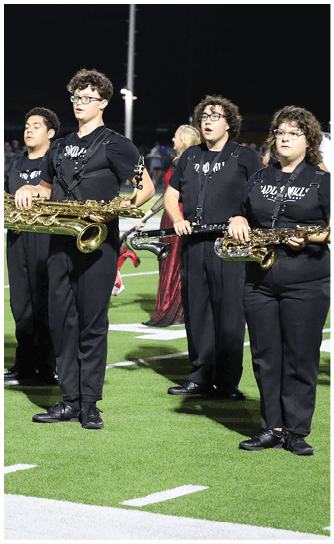 Two people dressed in black marching band uniforms stand on a football field, one holding a large brass saxophone. Descripci n generada con IA