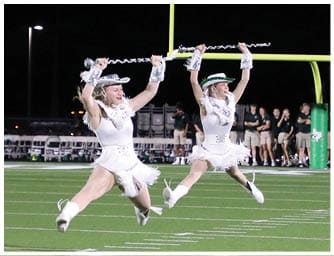 Two people in white sequined costumes and hats are mid-jump performing a baton twirling routine on a football field at night with a group of people standing near the goalpost in the background. Descripci n generada con IA