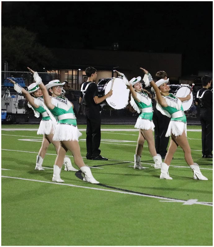 People in matching green and white uniforms perform a coordinated dance routine on a football field while others play drums in the background. Descripci n generada con IA