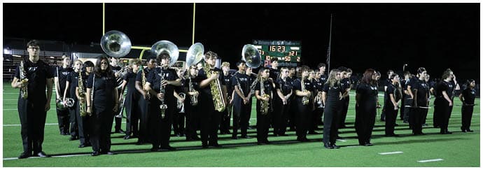 A group of people dressed in black uniforms are standing on a football field holding musical instruments as part of a marching band performance at night. Descripci n generada con IA