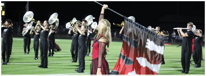 A person with long hair wearing a red and black outfit is holding and waving a large flag while a group of people in black uniforms play brass instruments on a field at night. Descripci n generada con IA