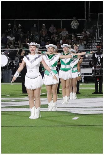 A group of people in matching white and green uniforms perform a synchronized routine on a football field at night with a marching band playing behind them. Descripci n generada con IA