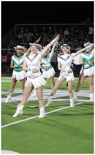 A group of adults dressed in white and green dance uniforms with hats are performing a synchronized high-kick routine on a football field at night. Descripci n generada con IA