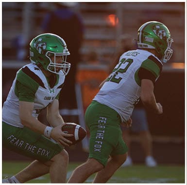 Two people wearing green helmets and white sports uniforms are engaged in a football game on a field. Descripci n generada con IA