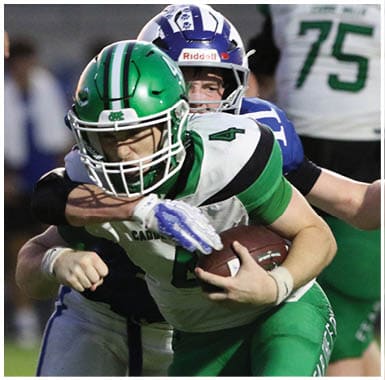 Four people in football uniforms are engaged in a play on a field, with one person in green lying on the ground while others in green and white surround them. Descripci n generada con IA