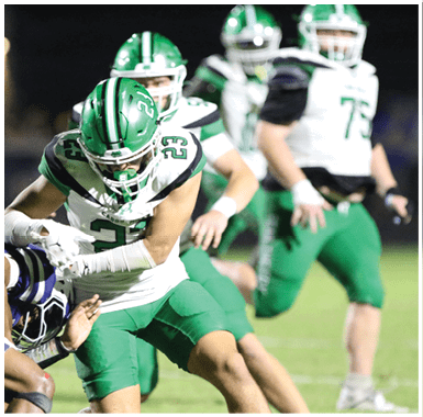 A person in a green football uniform is holding the ball while being tackled by two people in white and black uniforms on a football field. Descripci n generada con IA
