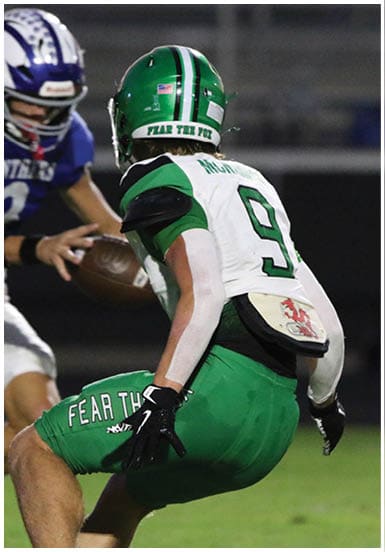 A person in a green football uniform with the number 34 is jumping over another person in a white football uniform during a nighttime game. Descripci n generada con IA