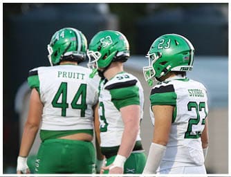 Three people wearing green and white football uniforms and helmets stand on a field with a blurred background. Descripci n generada con IA