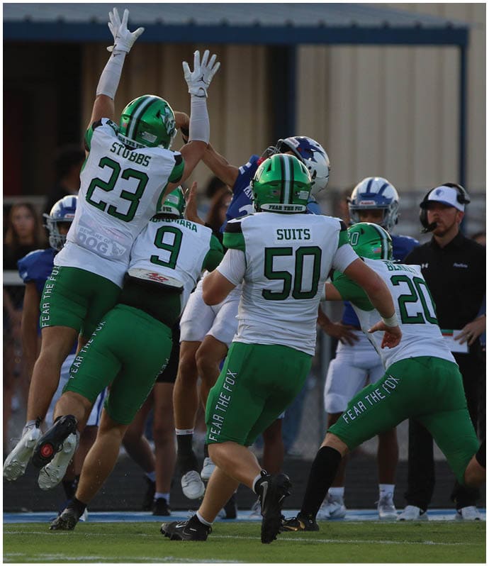 Two people in green football uniforms and helmets jump in celebration on a football field while others stand in the background near a golf cart and band members. Descripci n generada con IA
