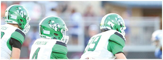 Two people in green football uniforms are running on a field during a game with a blurred background of spectators and equipment. Descripci n generada con IA