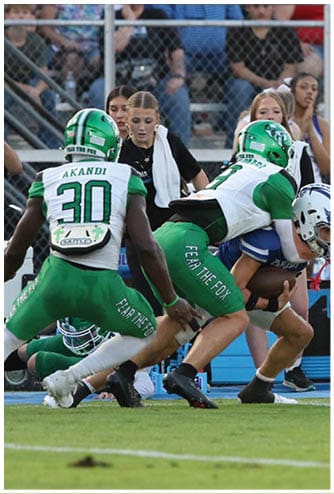 A person in green football gear and helmet is catching a football on a field with a blurred background of a fence, vehicles, and a sign. Descripci n generada con IA