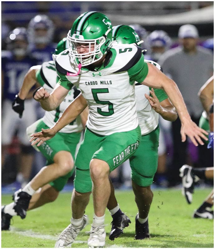 A group of football players in green uniforms are gathered on the field near the end zone while a referee stands in the foreground and the scoreboard shows the home team leading. Descripci n generada con IA
