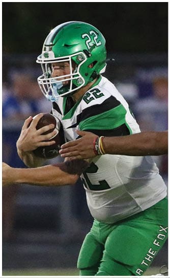 Two people in green football uniforms are on a field with one holding a football and the other preparing to receive it while players in white and black uniforms stand in the background. Descripci n generada con IA