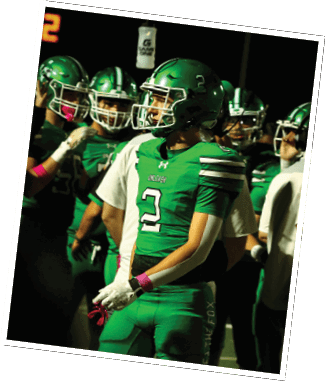 Two people in green and white football uniforms with helmets are on a field, one flexing their arms while the other stands nearby. Descripci n generada con IA