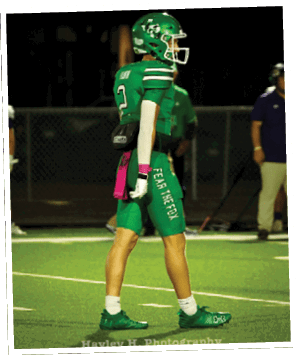 A person in a green football uniform is holding a football and appears to be preparing to throw it on a football field. Descripci n generada con IA