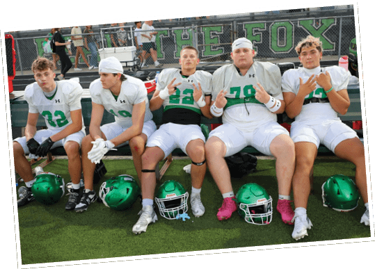 Three people in football uniforms sit on a bench on a field with helmets on the ground in front of them, making hand gestures and looking at the camera. Descripci n generada con IA