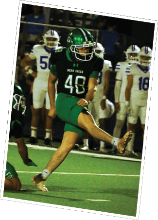 A person in a green and black football uniform is kicking a ball while another person in a similar uniform is crouched nearby, with people in white uniforms standing in the background. Descripci n generada con IA