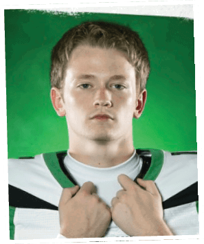 A person in a green football uniform is holding a football and appears to be preparing to throw it on a football field. Descripci n generada con IA