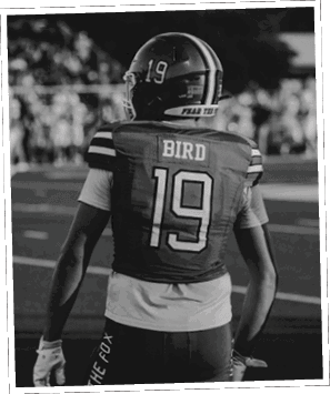 A person wearing a white and green football uniform with the name “Caddo Wolves" is smiling and looking at the camera with other players in green helmets in the background. Descripci n generada con IA