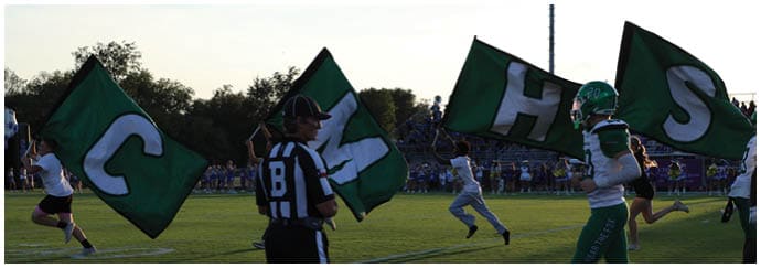 People running on a sports field holding large green flags with white letters while a referee in black and white stripes stands nearby. Descripci n generada con IA