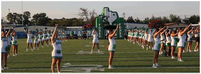 People in cheerleading uniforms are performing a routine on a football field with a scoreboard and inflatable decorations in the background. Descripci n generada con IA