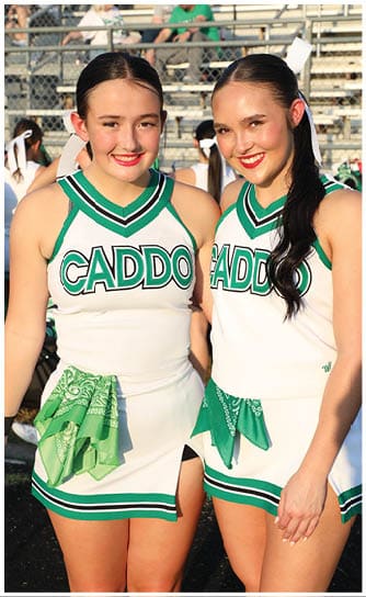 Two people wearing green and white cheerleading uniforms smile while standing close together in front of bleachers. Descripci n generada con IA