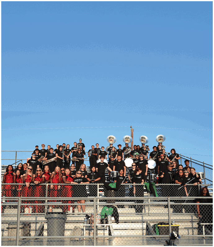 A large group of people wearing black and red uniforms are standing and sitting on bleachers outdoors under a clear blue sky, with some holding musical instruments. Descripci n generada con IA