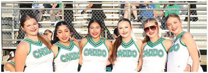Six adults wearing matching cheerleading uniforms with the word Caddo stand closely together smiling outdoors near a chain-link fence. Descripci n generada con IA