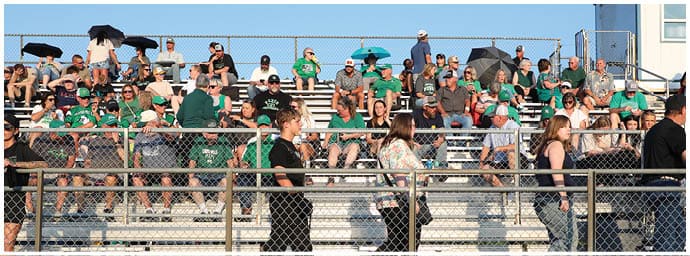 Adults wearing green shirts and casual clothing are seated and standing on metal bleachers outdoors, some holding umbrellas for shade. Descripci n generada con IA