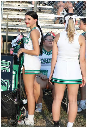 People in cheerleading uniforms stand near a cart holding signs and pom-poms on a sports field. Descripci n generada con IA