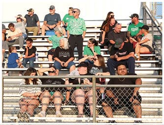 A diverse group of people wearing casual clothing and sunglasses are seated on bleachers outdoors, some shading their eyes from the sun while watching an event. Descripci n generada con IA