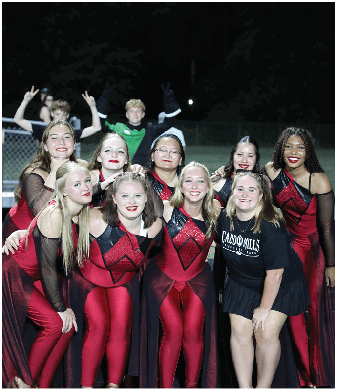 A group of people dressed in matching red and black performance outfits pose together on a track field at night with two people in the background making peace signs. Descripci n generada con IA