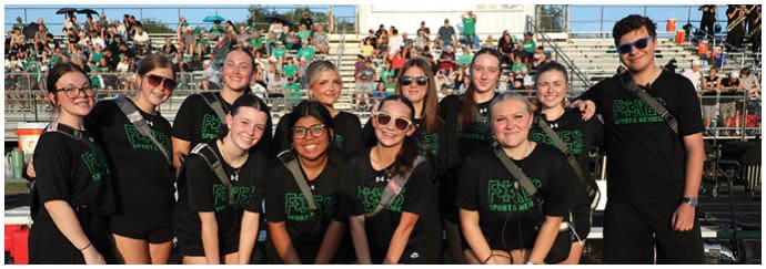A group of adults wearing matching black shirts with green text are posing together outdoors in front of bleachers filled with people. Descripci n generada con IA