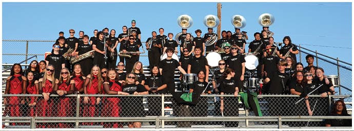 A large group of people wearing coordinated black and red outfits are seated and standing on bleachers, holding various musical instruments and smiling outdoors under a clear sky. Descripci n generada con IA