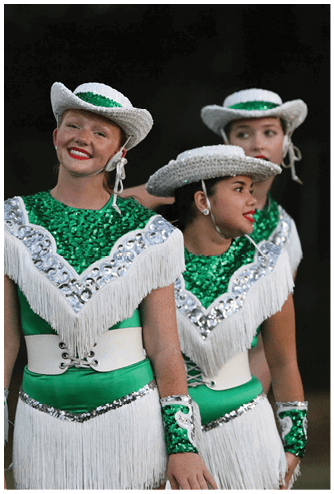A person wearing a green and white sequined dance costume with fringe and a matching white cowboy hat is smiling outdoors. Descripci n generada con IA