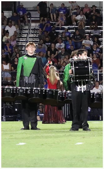 Five people in green and black uniforms stand on a football field holding percussion instruments with a crowd seated in bleachers behind them. Descripci n generada con IA
