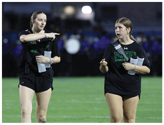 Two adults wearing black sports shirts and shorts are walking and talking on a grassy field with a blurred background of people and drums. Descripci n generada con IA