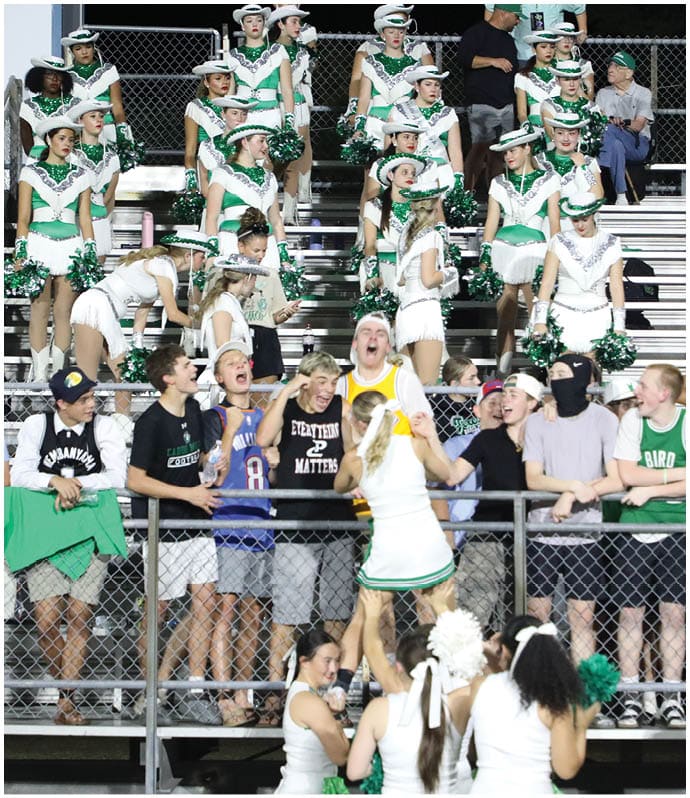 A group of people in casual clothing and cheerleaders in green and white uniforms are gathered in bleachers at an outdoor event, with some holding pom-poms and others engaging in conversation. Descripci n generada con IA