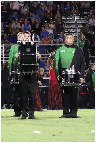 People wearing black pants and green tops are standing on a field holding drums, with others in red dresses visible in the background. Descripci n generada con IA