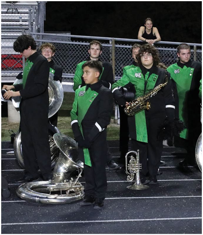 People in green and black marching band uniforms stand on a track at night with musical instruments while one person leans on a fence behind them smiling. Descripci n generada con IA