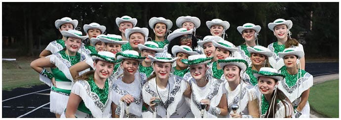 A group of people dressed in white and green dance uniforms with hats are smiling and posing together on an outdoor track. Descripci n generada con IA