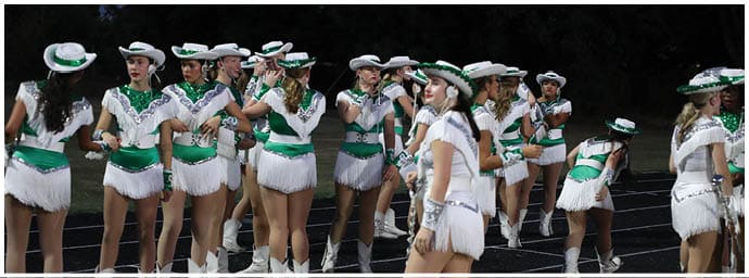 A group of people dressed in white and green fringed outfits with hats are standing and interacting on a track field at night. Descripci n generada con IA