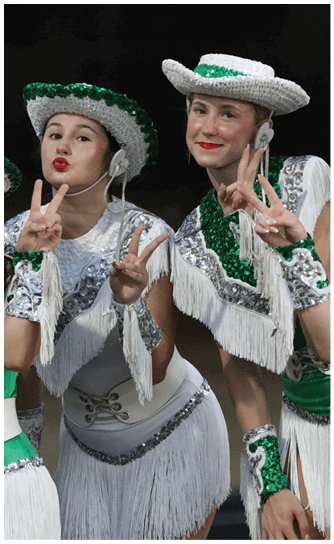 Two people wearing sequined green and white costumes and hats are smiling, with one person making a peace sign gesture. Descripci n generada con IA