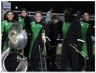 People in green and black marching band uniforms stand on a track holding various brass instruments at night with bleachers and a fence in the background. Descripci n generada con IA