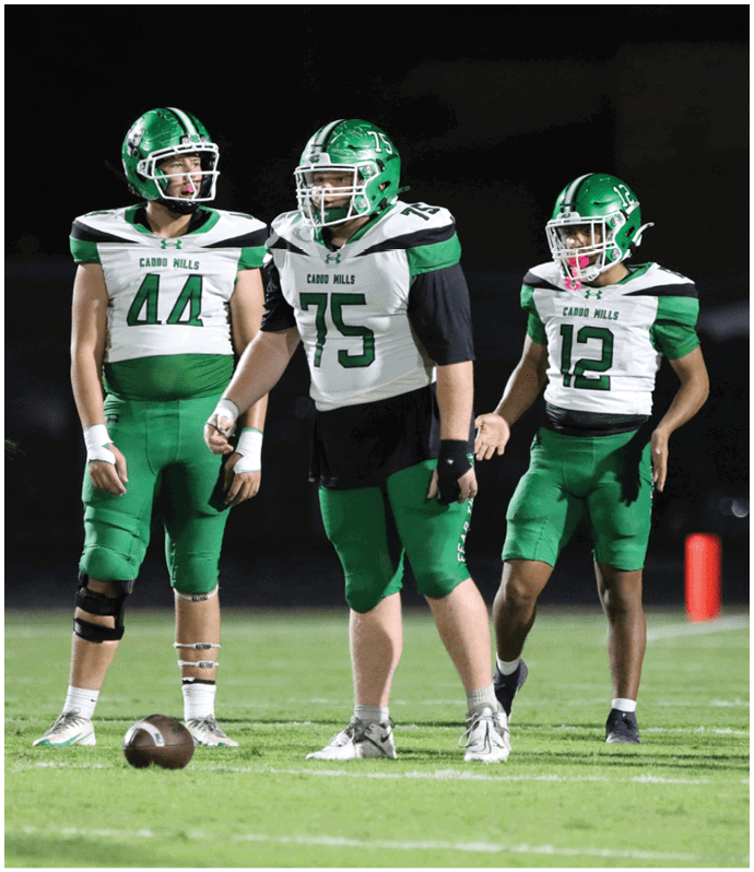 A group of football players in green uniforms are gathered on the field near the end zone while a referee stands in the foreground and the scoreboard shows the home team leading. Descripci n generada con IA