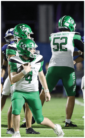 Two people in green football uniforms are on a field with one holding a football and the other preparing to receive it while players in white and black uniforms stand in the background. Descripci n generada con IA
