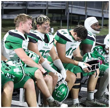 Several people in green and white football uniforms are sitting on a bench on the sidelines of a field, some holding helmets and appearing focused or resting. Descripci n generada con IA