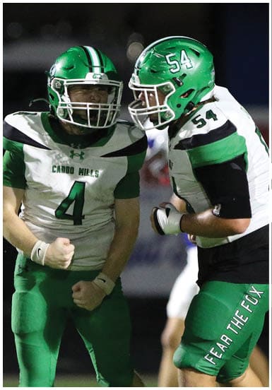 A person in a green football uniform with the number 34 is jumping over another person in a white football uniform during a nighttime game. Descripci n generada con IA